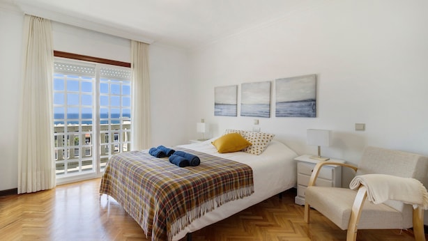 Un dormitorio con cama, una silla, una mesita de noche y vista al mar a través de una ventana.