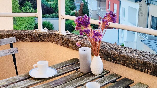 A balcony with a wooden table set for two, a white vase with purple flowers, and a white candle. - 2 bedrooms in the heart of city center (Luxembourg City)