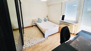 A bedroom with a bed, bedside table, chair, radiator, window with blinds, and a hanging light fixture. - 2 bedrooms in the heart of city center (Luxembourg City)