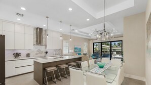 A modern kitchen with a central island, bar stools, and a dining area with a glass table. - Villa Del Mare Beach 4 Ensuite Retreat + Heated Pool – Special Offer! (Naples)