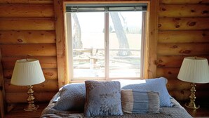 A log cabin bedroom with a bed, two bedside lamps, and a window with a view of trees and a wooden deck.