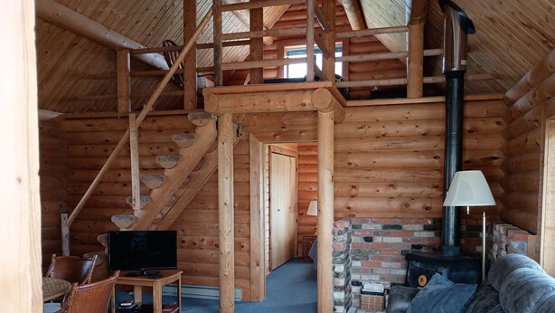 A log cabin interior with a wooden staircase, a brick fireplace, a sofa, and a television.