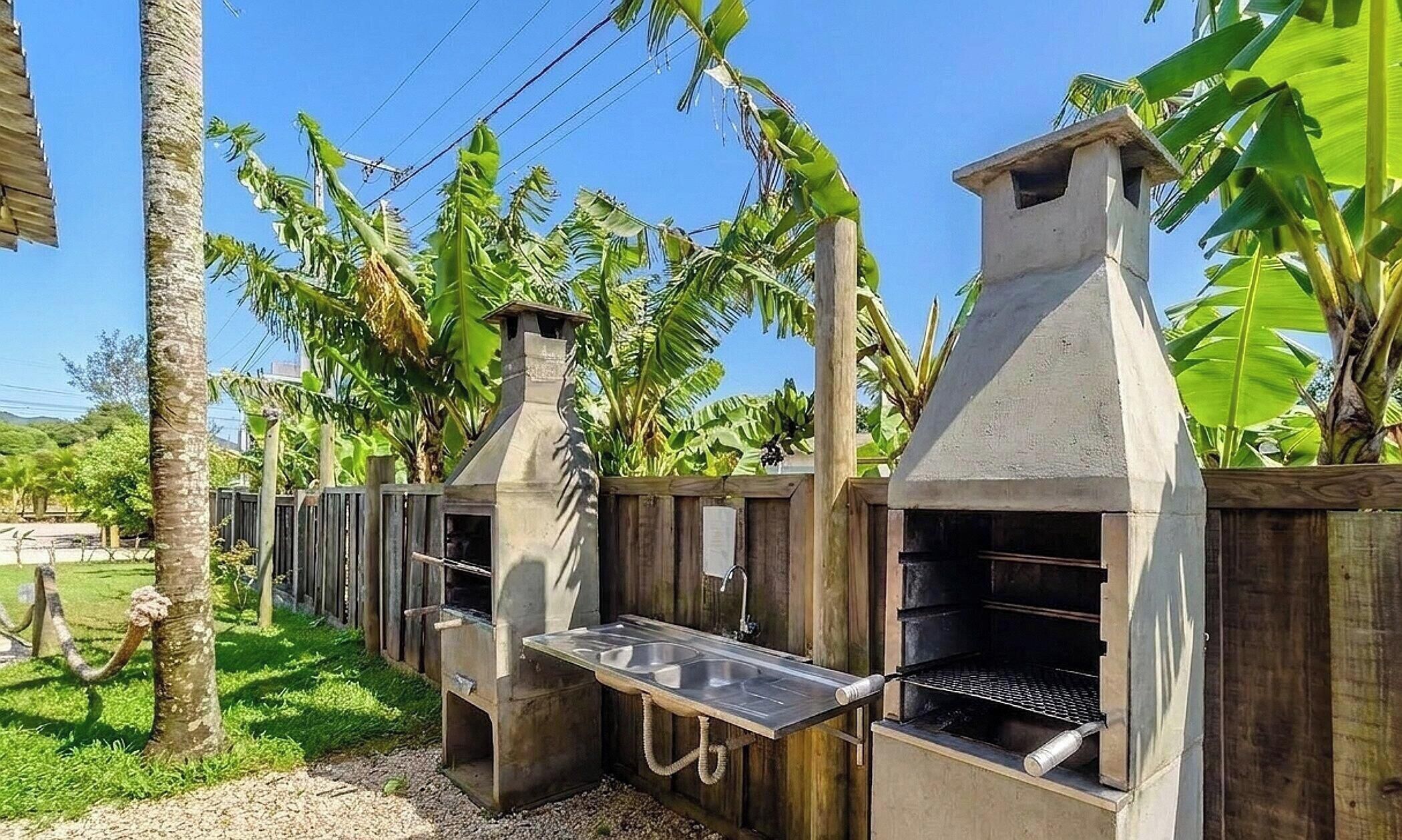 An outdoor kitchen with a grill and a sink, surrounded by wooden fencing and banana trees.