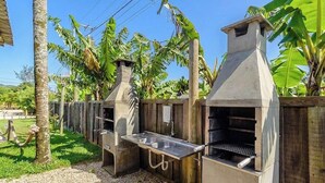 Una cucina esterna con una griglia e un lavandino, circondata da un recinto in legno e vegetazione rigogliosa.