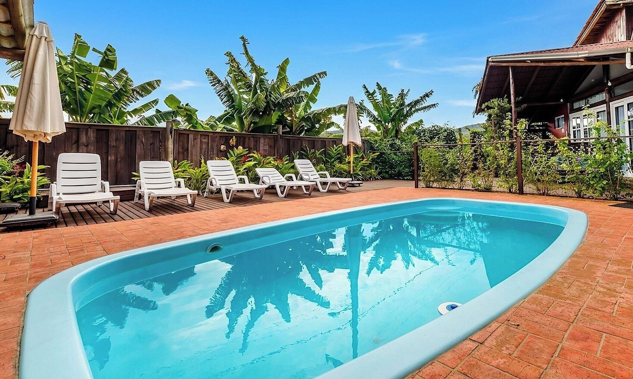A rectangular swimming pool with white lounge chairs and umbrellas, surrounded by a wooden fence and lush greenery.