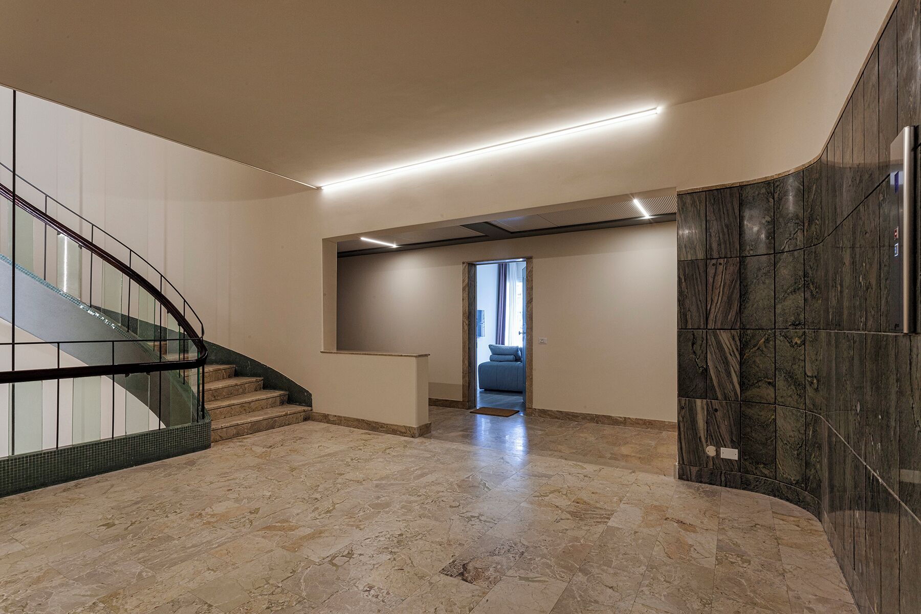 A spacious interior with a curved staircase, a marble floor, and a view into a bedroom through a doorway.