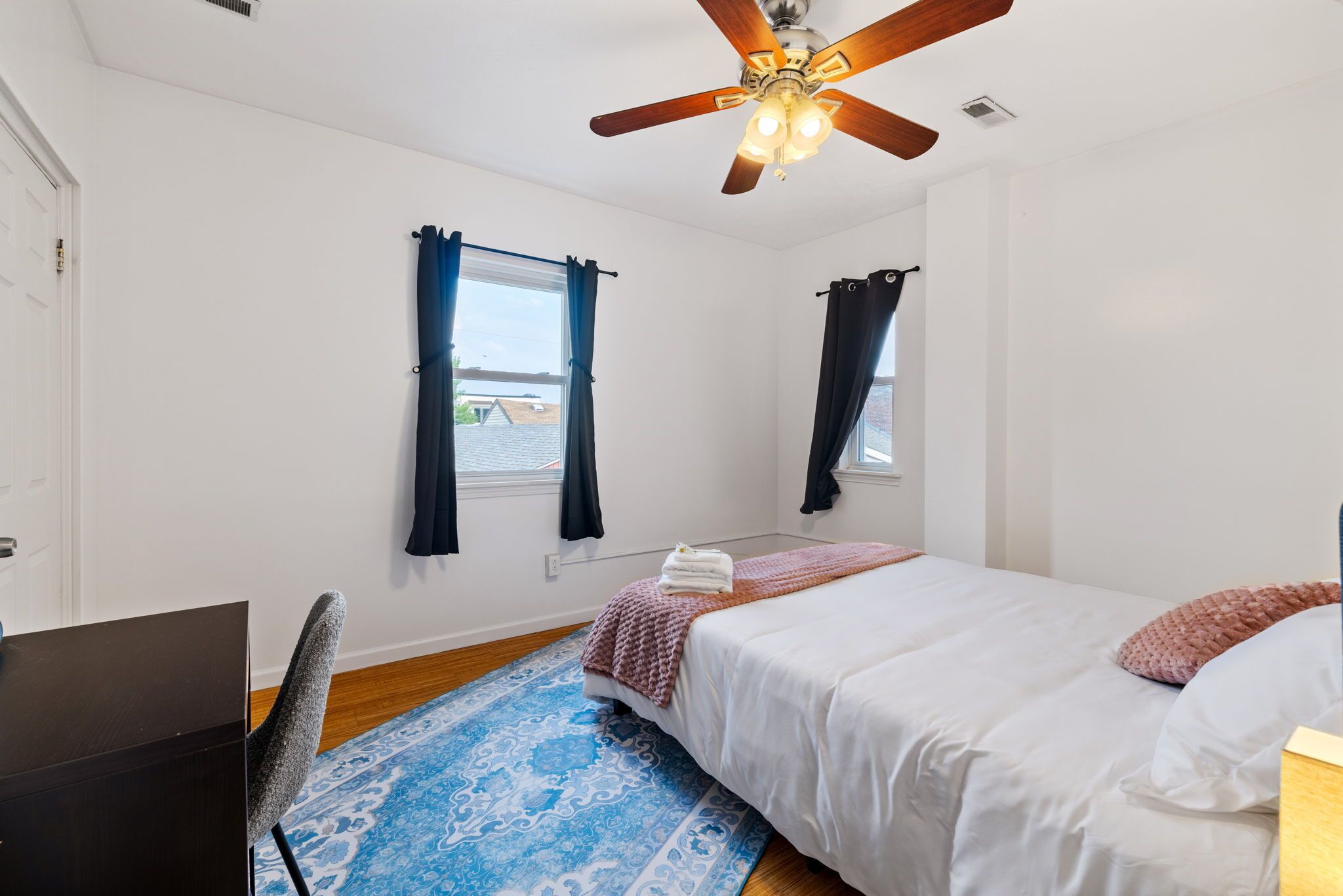 Ein Schlafzimmer mit einem Bett, einem Schreibtisch, einem Sessel, einem Deckenventilator und zwei Fenstern mit Vorhängen.