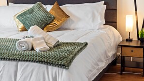 Een net opgemaakt bed met witte lakens, een groene gebreide plaid en een opgerolde witte handdoek. Er liggen twee kussens op, één groen en één goudkleurig. Een klein nachtkastje met een lamp en een plantje is zichtbaar.