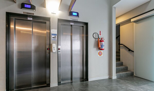 Zwei Aufzugtüren mit digitalen Anzeigen, ein Feuerlöscher und eine Treppe.