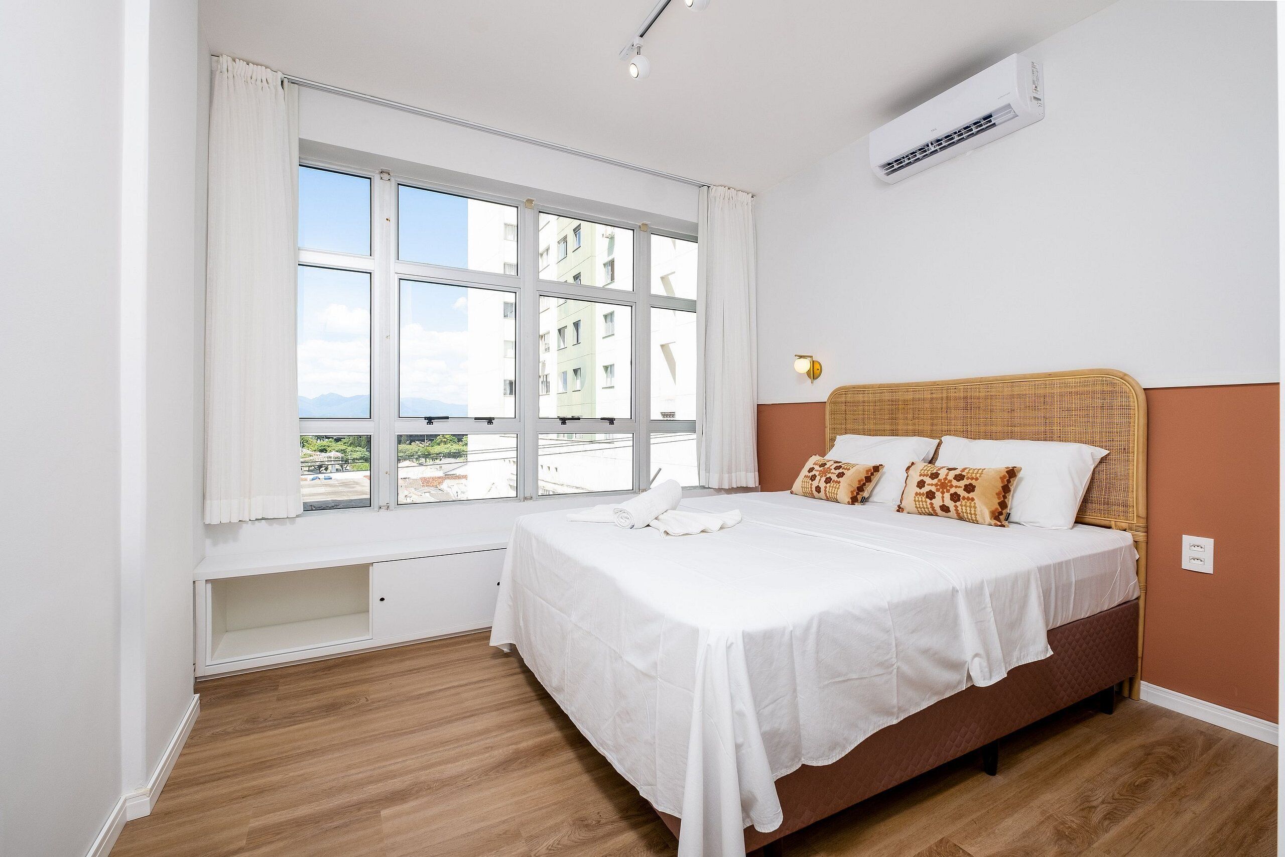 Un dormitorio con un ventanal grande, una cama con ropa de cama blanca, un cabecero con diseño tejido y un equipo de aire acondicionado en la pared.