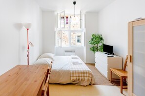 Une chambre à coucher comprenant un lit, une table de chevet, un bureau, une chaise, un téléviseur et une fenêtre donnant sur un bâtiment en pierre.