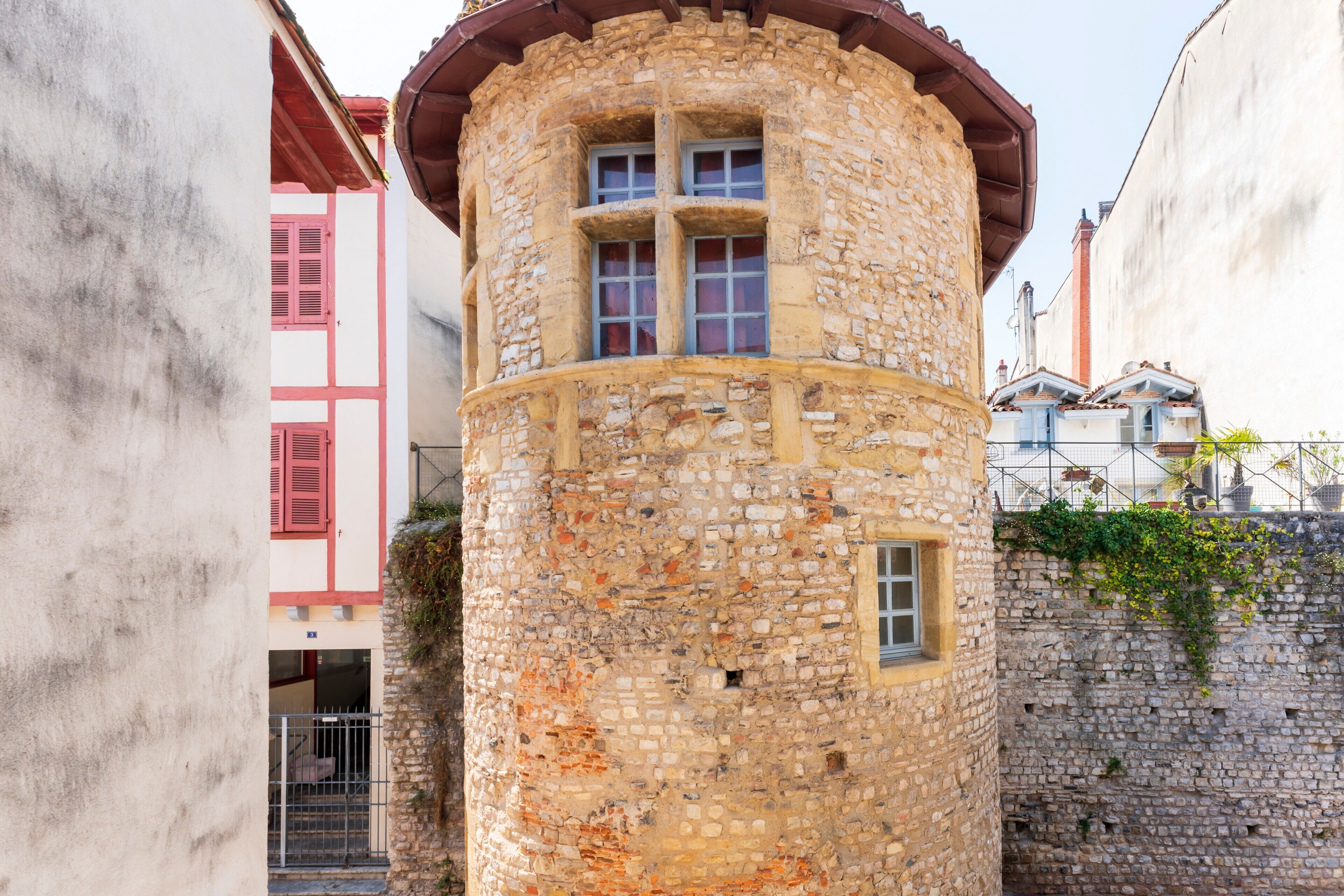 A stone tower with windows and a red roof, adjacent to a building with pink shutters.