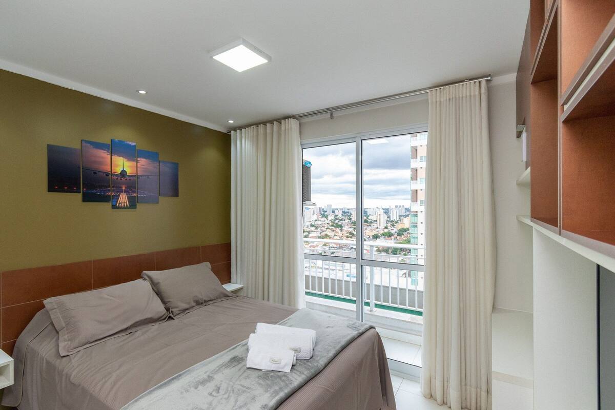 A hotel room with a bed, a city view, a painting on the wall, and a window with curtains.
