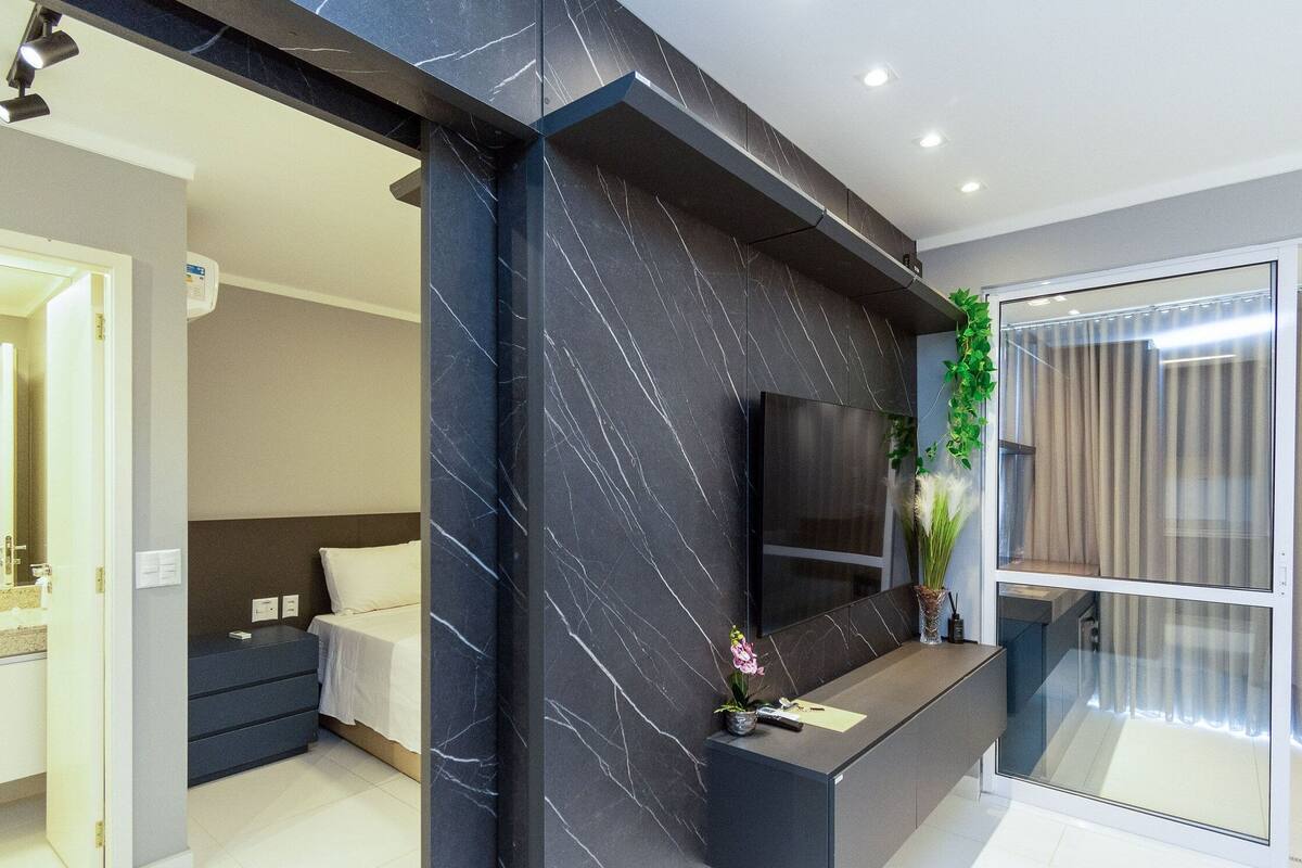 A modern hotel room with a dark marble accent wall, a bed, a bedside table, a TV, and a glass partition.