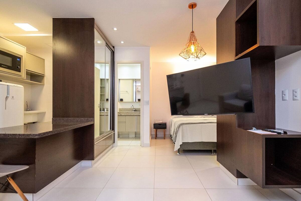A modern hotel room with a kitchenette, a large flat-screen TV, a bed, and a chandelier.