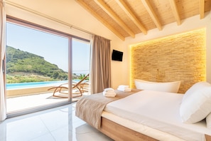 A bedroom with a bed, a TV, a stone wall, and a view of a beach and mountains.