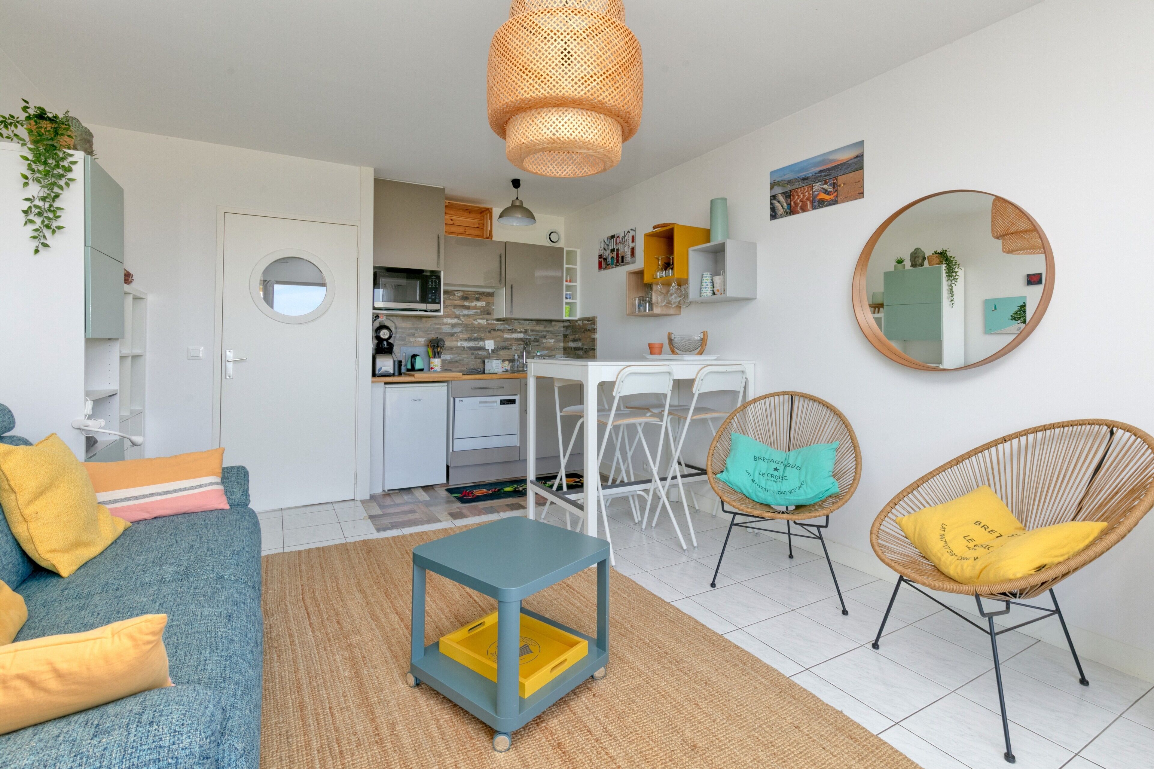 A modern living room with a sofa, coffee table, and chairs, a kitchen area with a dining table, and a round mirror on the wall.