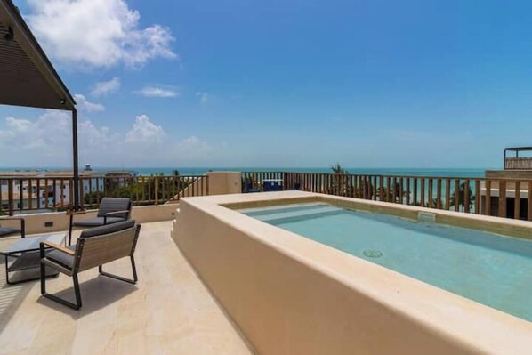 A rooftop pool with a clear view of the ocean, surrounded by lounge chairs and a covered seating area.