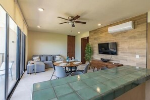 A modern living room with a dining area, a ceiling fan, a flat-screen TV mounted on the wall, and a large window providing natural light.