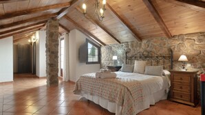 Une chambre avec un mur en pierre, des poutres en bois au plafond, un lit recouvert d’une couvre-lit à motifs, une table de chevet avec une lampe et une commode.
