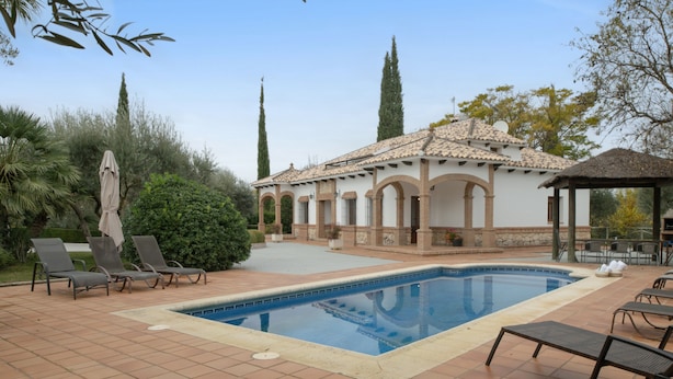 Une piscine avec des chaises longues, un espace de détente couvert et un bâtiment doté d ’arches et d’un toit en tuiles.
