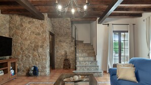 Un mur de pierre, un escalier, une table basse en verre, un canapé bleu et un téléviseur.