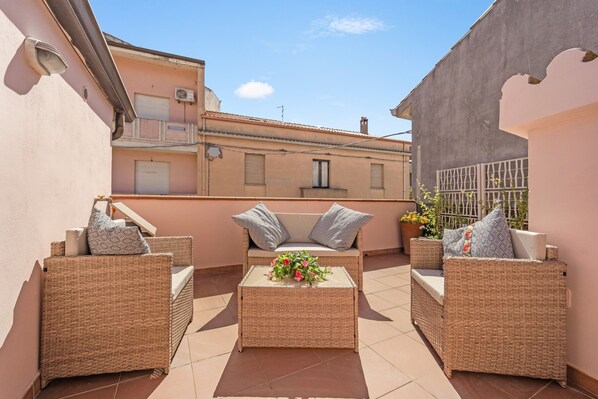 Un balcón con muebles de mimbre, una mesa pequeña con flores y vista a edificios.