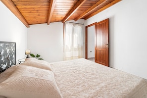 Un dormitorio con techo de madera, una cama con colcha estampada, una mesita de noche con lámpara y una puerta que da a otra habitación.