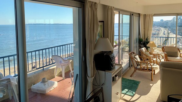 Een balkon met uitzicht op zee en stadsgezicht, voorzien van stoelen en een tafel, en een televisie op een standaard.