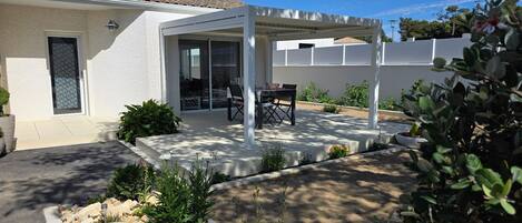 Een huis met een overdekte patio, een deur met een raam in mozaïek en een tuin met verschillende planten en een rotspartij.