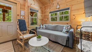 Interno accogliente di una baita con soffitto in legno, un divano, una poltrona, un tavolino e una finestra vista verde.