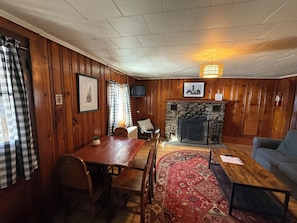 A cozy room with a wooden paneled wall, a fireplace, a dining table with chairs, a sofa, and a television mounted on the wall.
