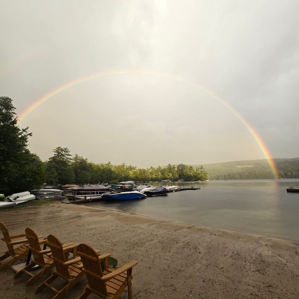 A lakeside scene with a rainbow, boats, and wooden chairs.