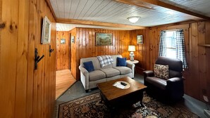Ein gemütlicher Raum mit Holzwänden und -decke, einer Couch, einem Sessel, einem kleinen Tisch und einer Lampe.