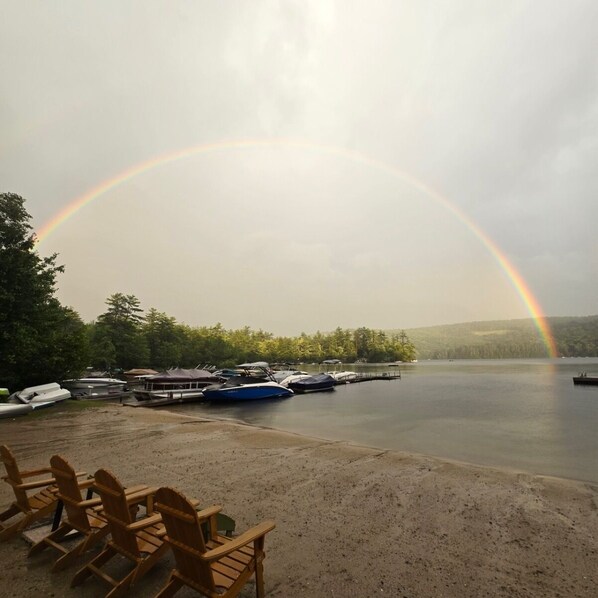 A lakeside scene with a rainbow, boats, and wooden chairs.