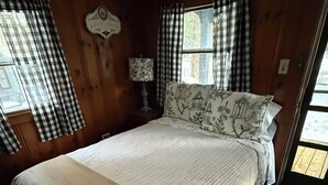 A bedroom with a large bed, checkered curtains, a nightstand with a lamp, and a window with a view of the outdoors. - Lakeside Cabin 7 with Kitchen (Holderness)