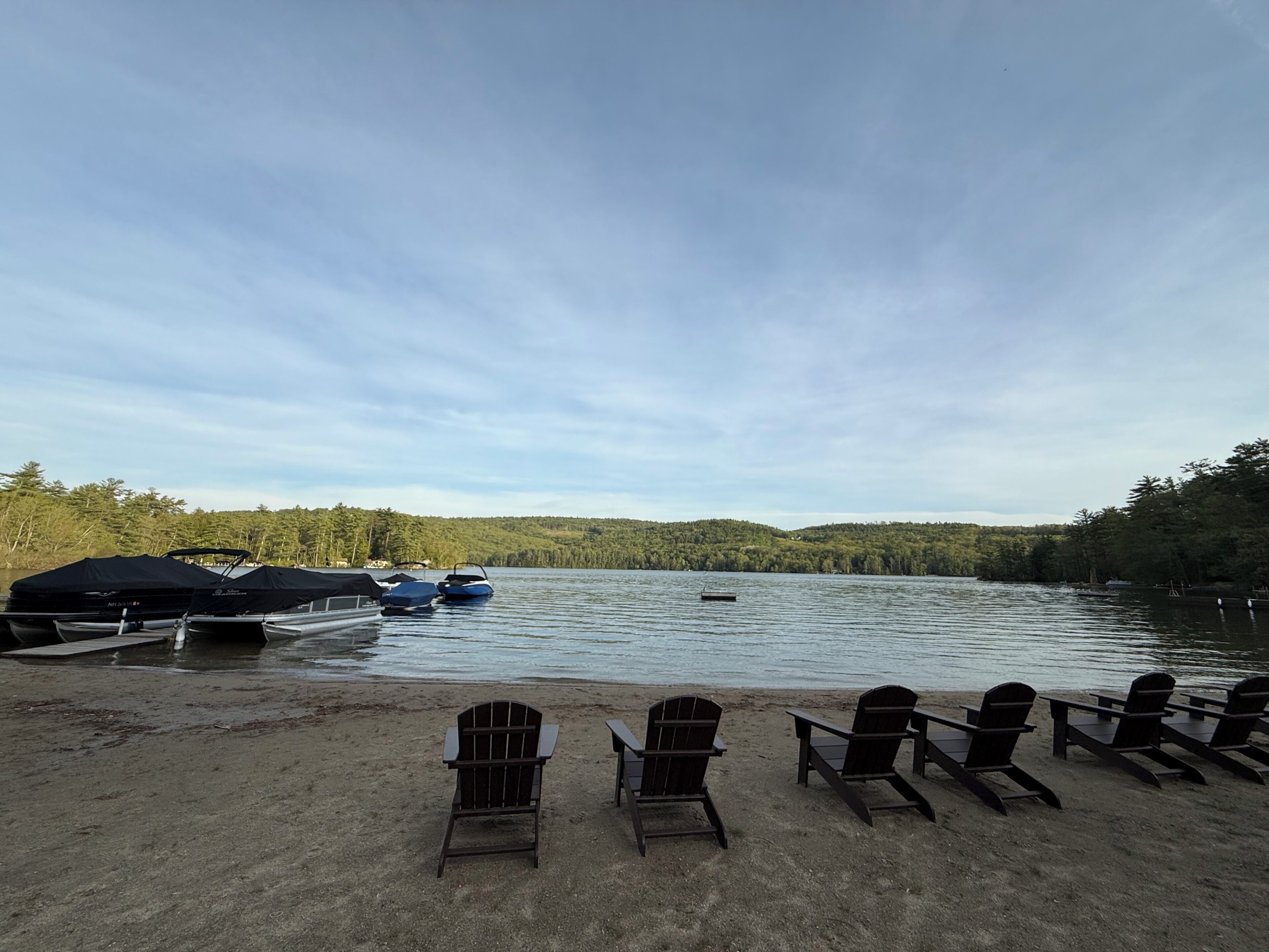 A lakeside view with docked boats, chairs, and a forested hill in the background.