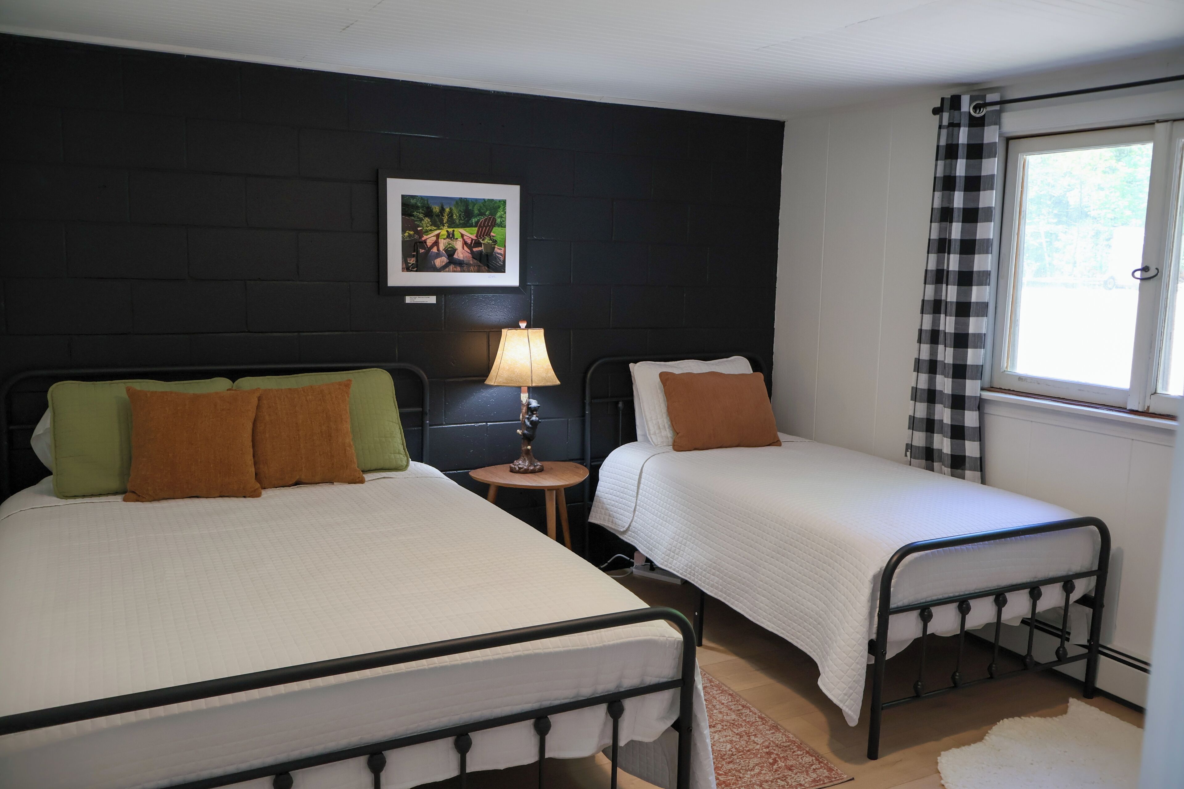 Une chambre avec deux lits, une table de chevet avec une lampe, un tableau au mur et une fenêtre avec un rideau à carreaux.