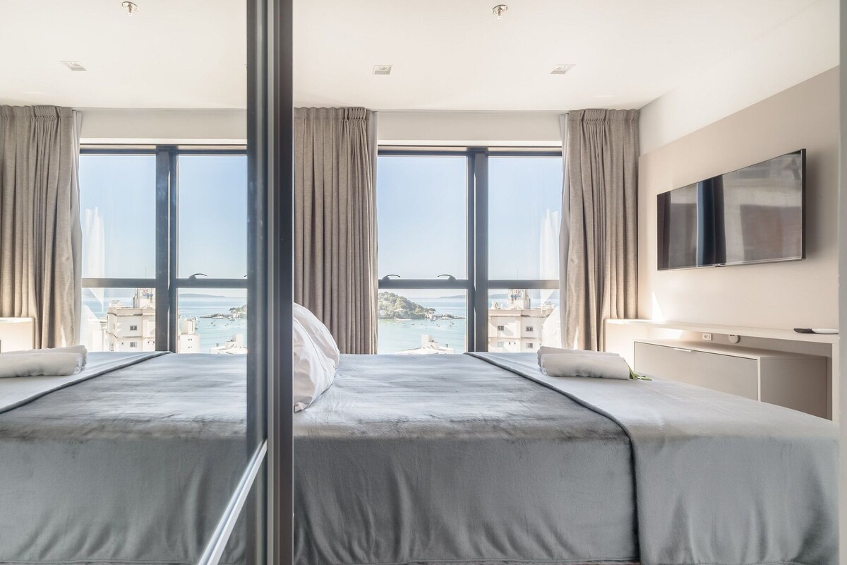 Une chambre d’hôtel avec un grand lit, deux écrans de télévision et une vue sur une ville côtière.