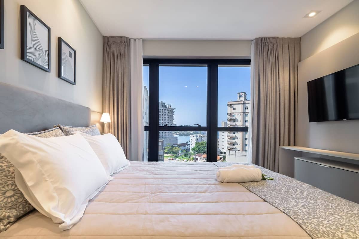 A hotel room with a large bed, a TV, a window with a city view, and framed artwork on the wall.