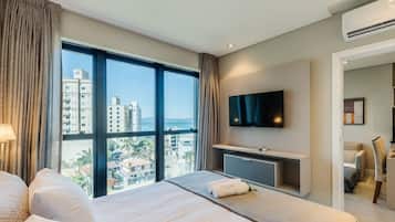 A hotel room with a large window, a bed, a desk, a flat-screen TV, and an air conditioning unit.