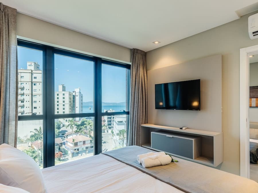 A hotel room with a large window, a bed, a desk, a flat-screen TV, and an air conditioning unit.