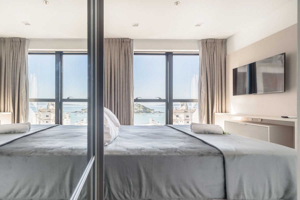 Habitación de hotel con una cama grande, dos televisores, vista al mar y un balcón.