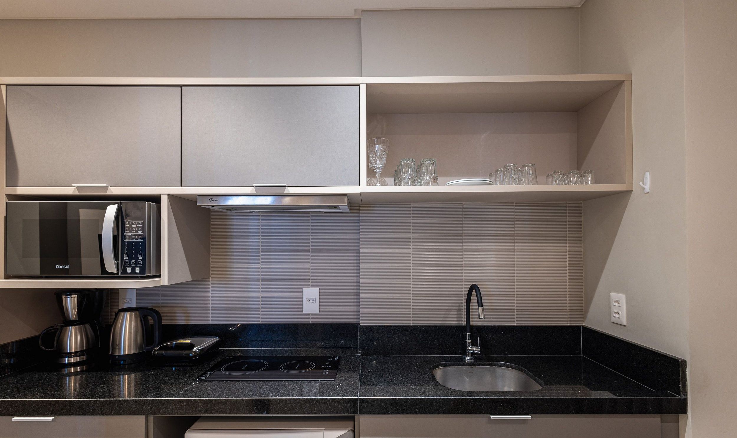 A compact kitchen with a microwave, kettle, and coffee maker on the countertop, a sink with a faucet, and a shelf with glassware.