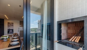 A modern kitchen with a built-in stove, a dining area with a table set for two, and a view of a cityscape through large windows. - MHFs Apartamentos com Vista Mar Itapema (Itapema)