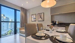 Un espace repas moderne avec une table dressée pour deux personnes, un canapé et une vue sur le paysage urbain.