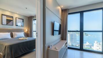 A modern hotel room with a bed, a TV mounted on the wall, a large window with a city view, and a small table with a remote control.