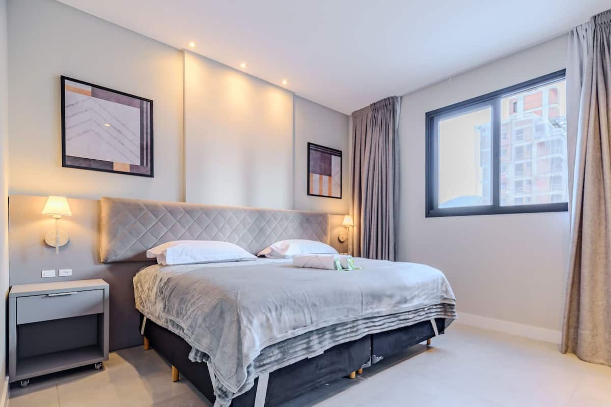 A modern hotel room with a large bed, bedside lamps, a nightstand, and a window with curtains.
