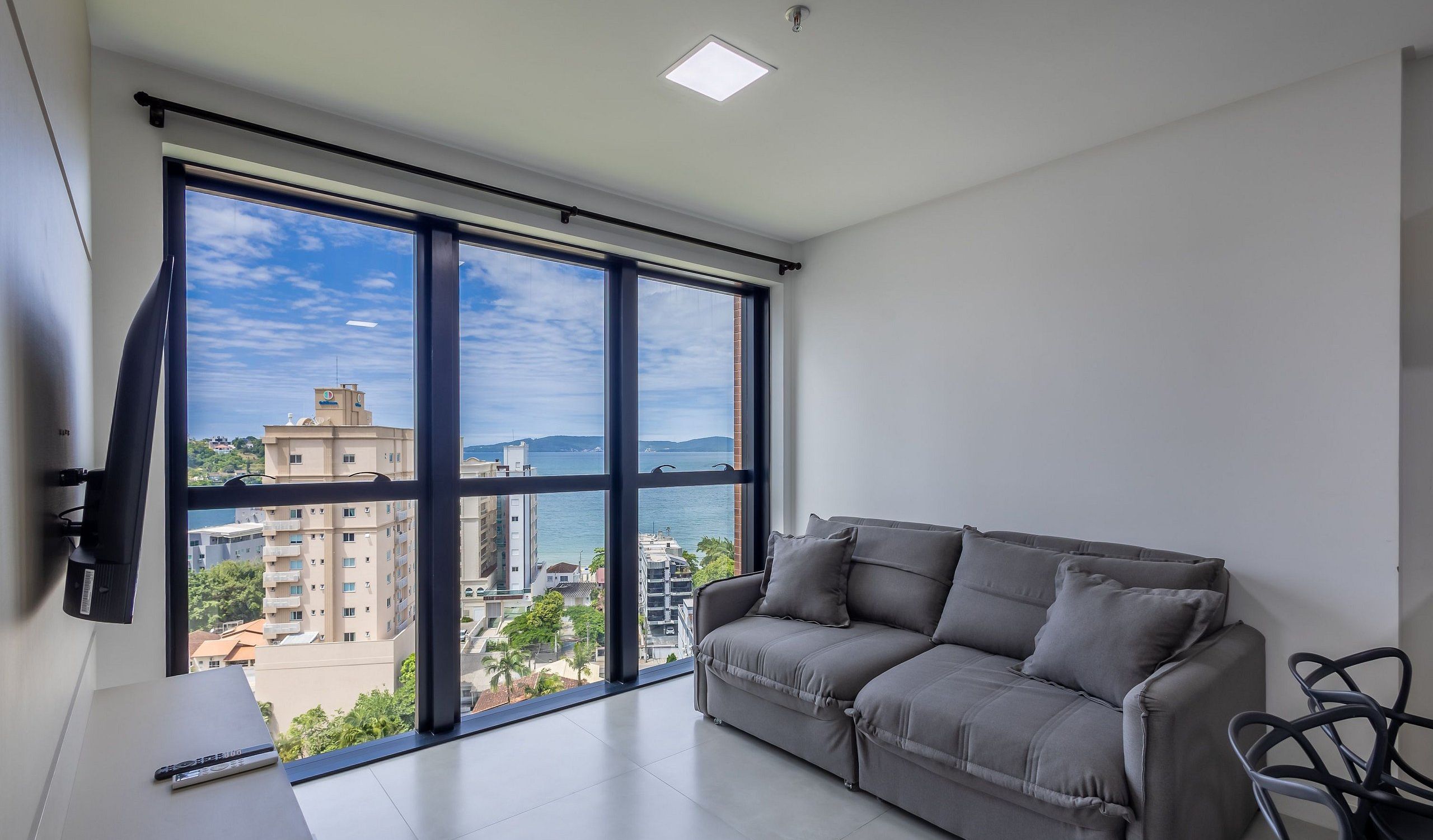 A modern living room with a grey sofa, a TV mounted on the wall, and a large window offering a view of buildings and a body of water.