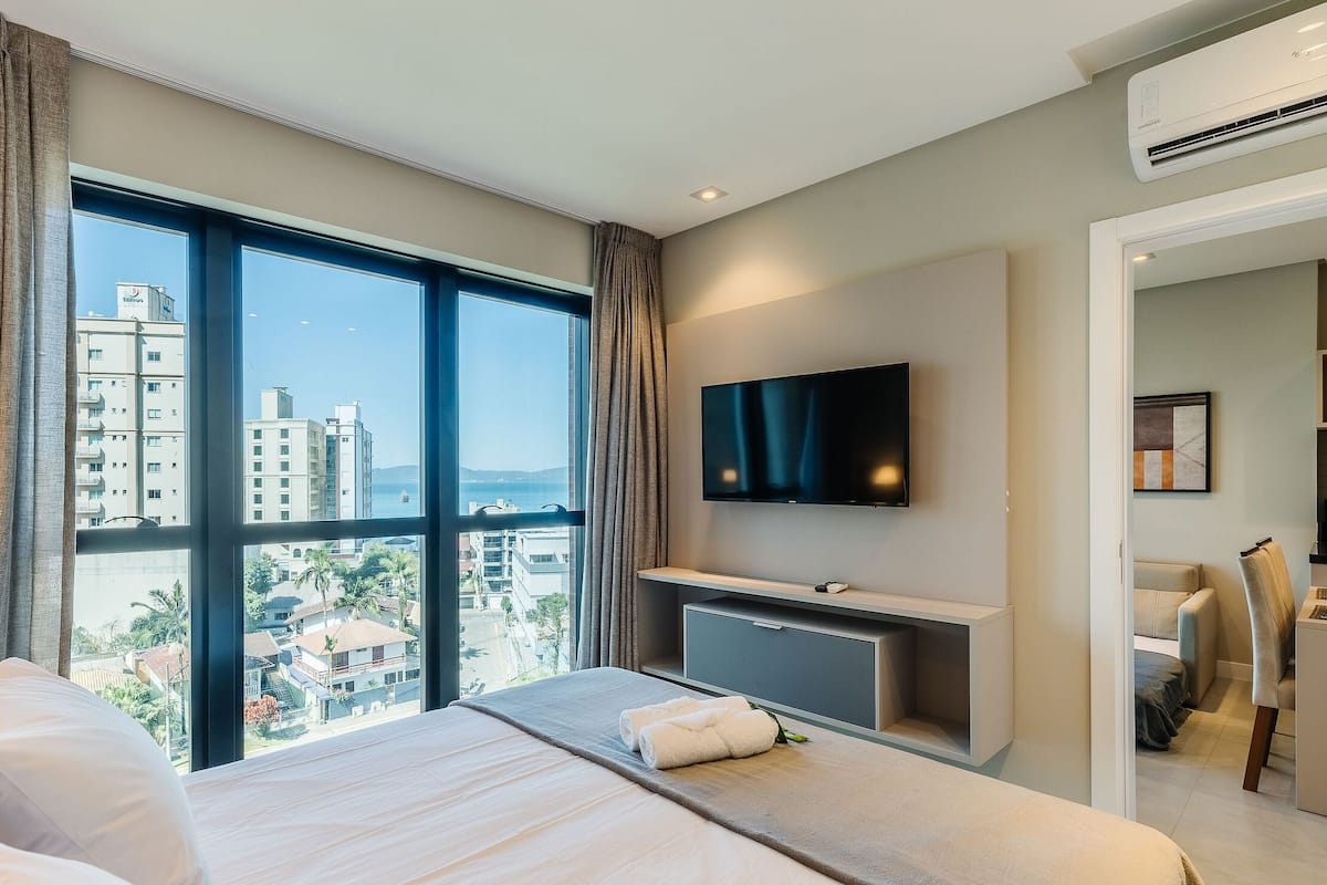 A hotel room with a large window, a flat-screen TV, a bed, a desk, and an air conditioning unit.